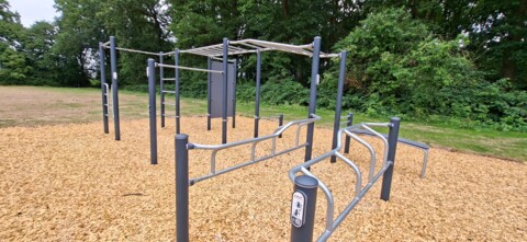 Das Foto zeigt die Callisthenics-Anlage in Münster-Angelmodde im Juli 2026 kurz nach der Fertigstellung, zu sehen sind in einer schrägen Ansicht die grauen Metallpfosten mit den angebauten silbernen Leitern und Bänken, der Boden ist mit Rindenmulch belegt, es sind keine Nutzerinnen zu sehen.  
