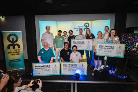 Das Bild zeigt alle Preisträger*innen nach Pitch und Prämierung auf der Bühne im Technologiehof Münster, links steht ein Banner mit dem Logo der Open Innovation City Münster auf türkis-gelbem Hintergrund. Es sind vier verschieden große Teams zu sehen, die jeweils einen riesigen Scheck über ihr Preisgeld halten, mittig vorne das Projekt 1buddy, das den ersten Platz erreicht hat.