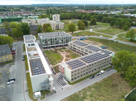 Das Bild zeigt eine Luftbildaufnahme aus geringer Höhe mit Blick über den vierseitigen Block des Grünen Weilers in Richtung Süden mit dem charakteristischen grünen Baukörper des vom-Stein-Gymnasiums im Mittelgrund links und den baumbestandenen Grünanlagen Grüner Trichter/Grüner Finger, die sich in Richtung Horizont ziehen. 
