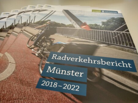 Einige Exemplare des neuen Radverkehrsberichts liegen aufgefächert auf einem Tisch. Das Titelbild zeigt Radfahrende auf einer Fahrradstraße, die auf einer neuen Kanalbrücke verläuft.