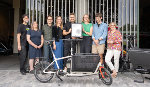 Das Bild zeigt 8 Personen nebeneinander zu einem Gruppenfoto aufgestellt vor der Fahrradwerkstatt des Wohnprojekts Grüner Weiler, von links nach rechts: Thorsten Liebold, Manuela Liebold, Ruben Grimm, Nicole de Vries, Christian Scheler vom Berliner Good Mobility Council mit der Preisurkunde, Cäcilia Stockmann, Felix Austen, Sabine Kittel, im Vordergrund und rechts halb verdeckt 2 Lastenräder