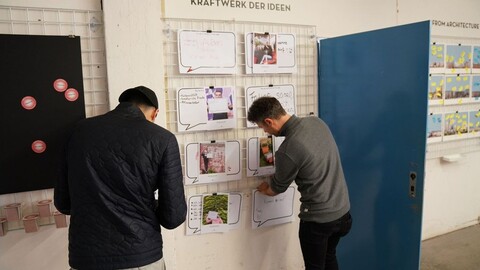 2 junge Männer befestigen in einem Workshop ihre Beiträge an einer Wand mit der Überschrift "Kraftwerk der Ideen"