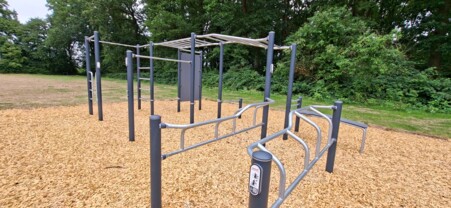 Das Foto zeigt die Callisthenics-Anlage in Münster-Angelmodde im Juli 2026 kurz nach der Fertigstellung, zu sehen sind in einer schrägen Ansicht die grauen Metallpfosten mit den angebauten silbernen Leitern und Bänken, der Boden ist mit Rindenmulch belegt, es sind keine Nutzerinnen zu sehen.  