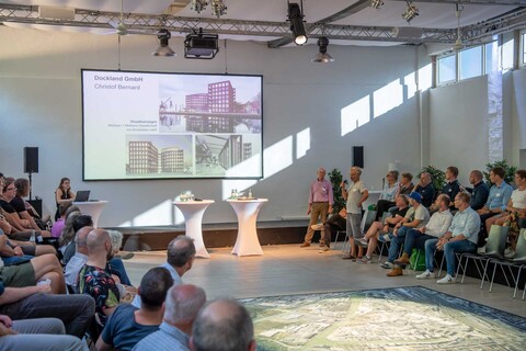 Christoph Bernard über den geplanten neuen Standort der Dockland GmbH, an die Wand projiziert eine Präsentationsfolie mit Architekturzeichnungen zum Projekt