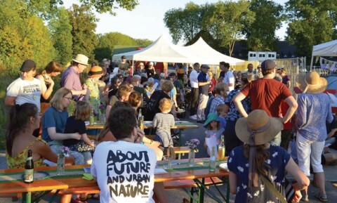 Das Bild zeigt zahlreiche Menschen bei einem Fest im Freien vor Büschen und Bäumen, teils stehend, teils auf Bierzeltganituren, in der Mitte ein weißer Zeltpavillon, im Hintergrund Baucontainer. Zu lesen ist die englischsprachige Aufschrift eines T-Shirts, in deutsch: "über die Zukunft und Stadtnatur" 