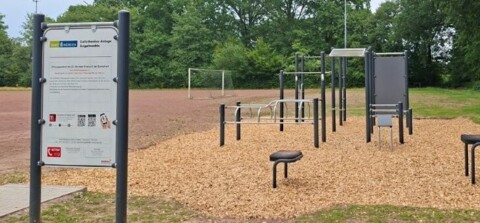 Das Foto zeigt die neue Calisthenics Anlage Angelmodde im Juli 2026 mit Hinweistafel zum Gebrauch und Beispielübungen unter dem Logo der Stadt Münster links im Bild neben einem frisch gepflasterten Fußweg, es sind keine Personen zu sehen, der Blick geht auf die Schmalseite der Anlage, links im Hintergrund ein Ascheplatz mit Fußballtor 