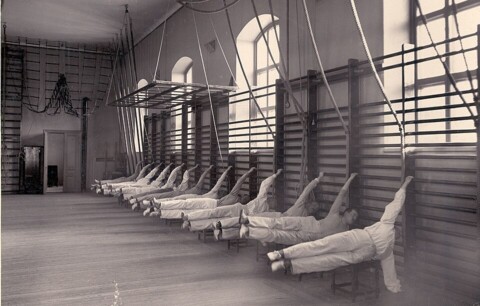 Eine historische Fotografie, die um das Jahr 1900 entstanden ist und ungefähr 10 Männer, vielleicht Soldaten oder Studenten, in zeitgenössischen hellen Trainingsanzügen in einer Turnhalle zeigt. Sie hängen alle parallel in einer langen Sprossenwand in der Haltung der "menschlichen Flagge" waagerecht in der Luft.